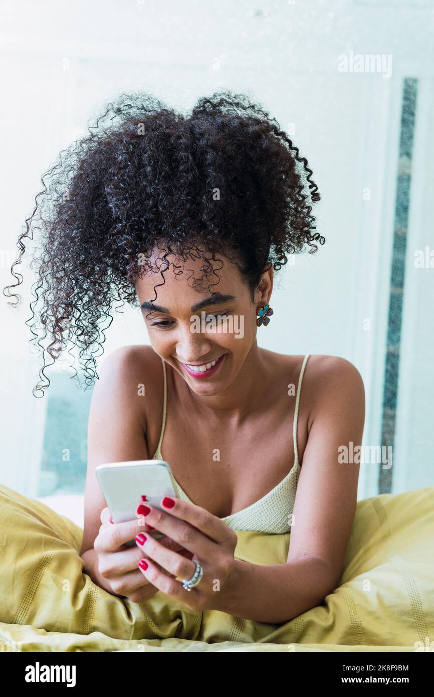 Happy curly-haired woman using smart phone at home Stock Photo
