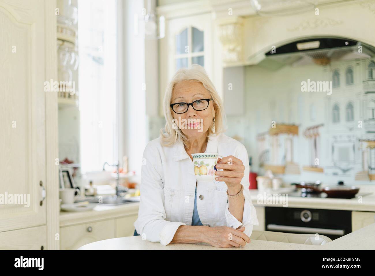Senior woman holding cup leaning on kitchen island at home Stock Photo