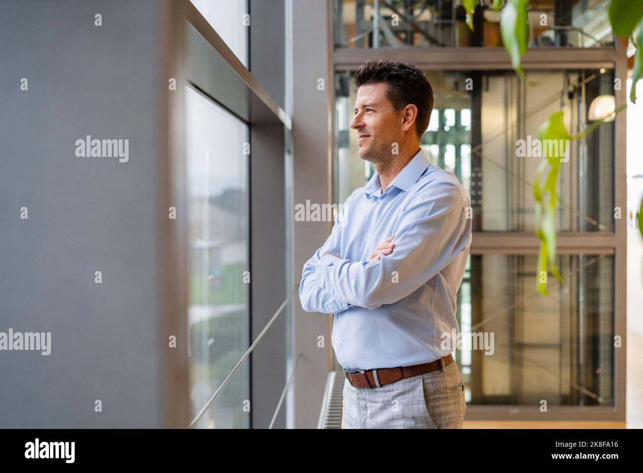 Smiling thoughtful businessman with arms crossed looking through window ...