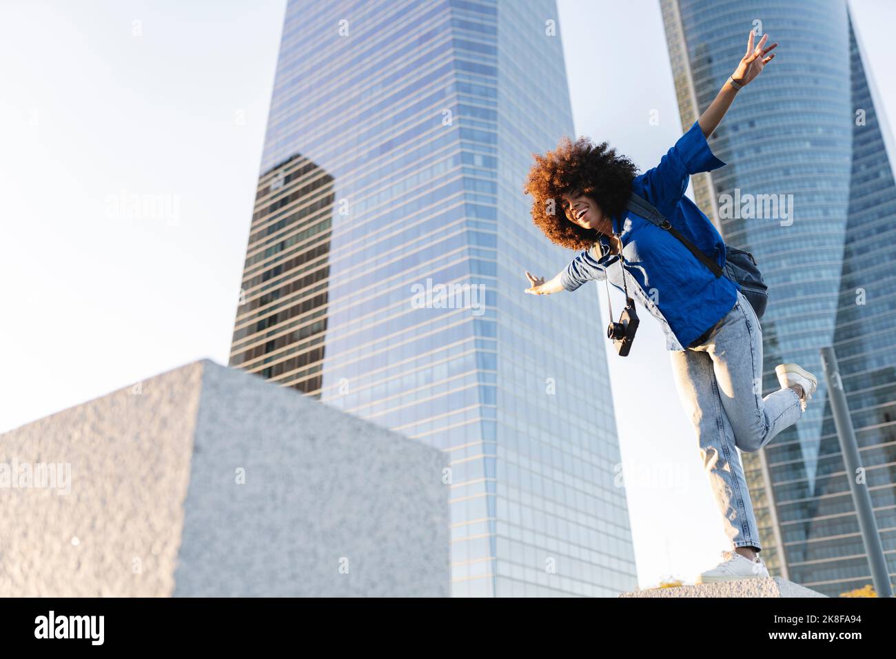 Smiling woman with arms outstretched standing on one leg in front of buildings Stock Photo