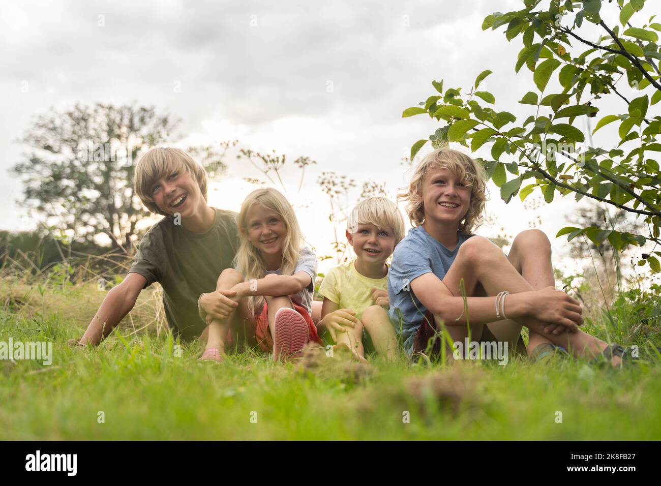 Smiling brothers and sister with blond hairs sitting on grass Stock Photo