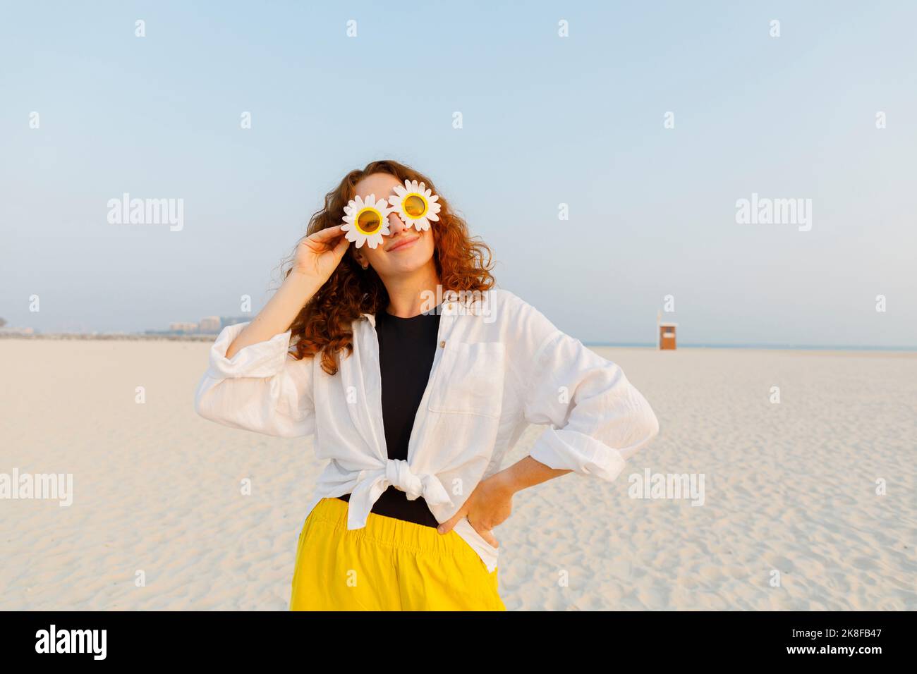 Smiling woman wearing sunflower sunglasses standing with hand on hip at beach Stock Photo