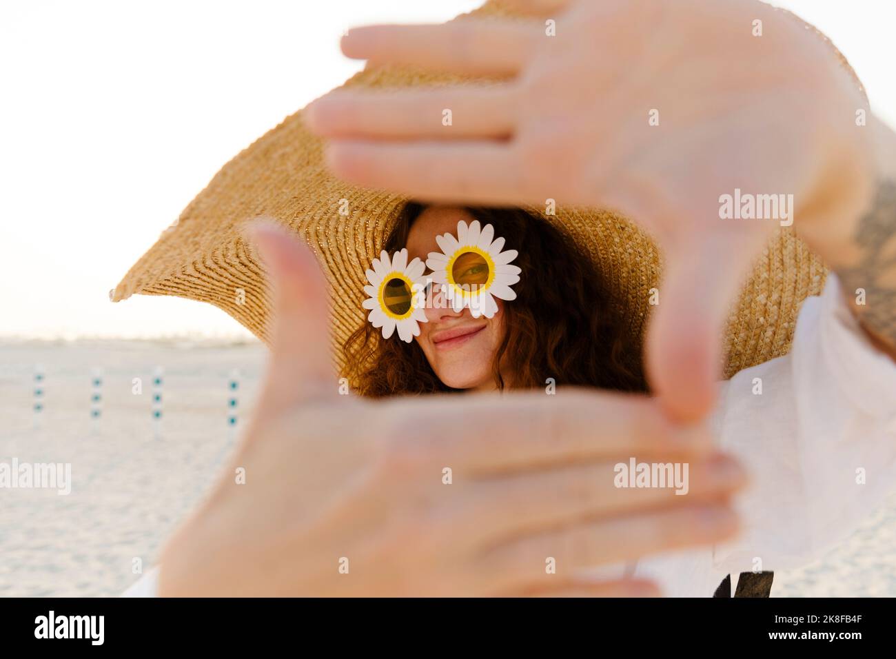 Smiling woman wearing sunflower sunglasses and oversized straw hat gesturing finger frame at beach Stock Photo