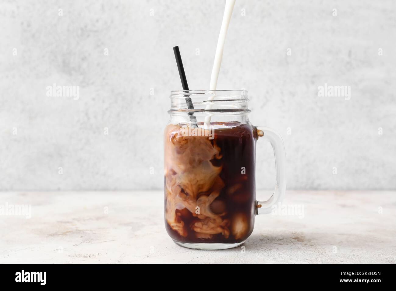 Pouring of milk into mason jar with delicious iced coffee on light