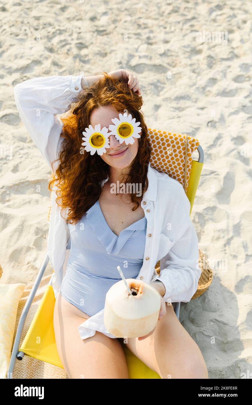Smiling woman wearing flower sunglasses sitting with coconut at beach Stock Photo