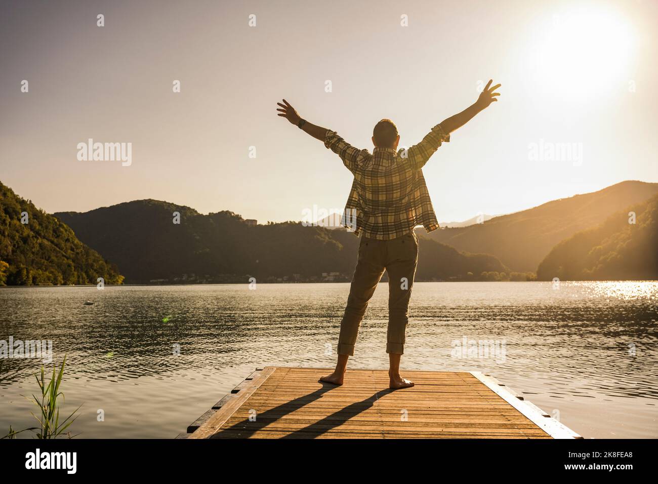 Mature man standing with arms raised on jetty by mountains Stock Photo