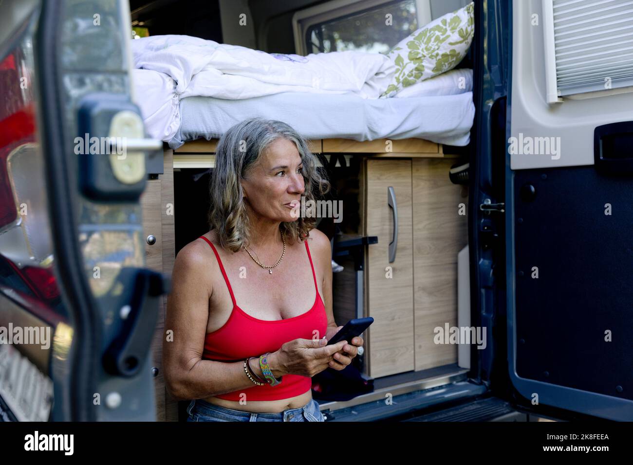 Smiling senior woman sitting with smart phone on bumper of camper van Stock Photo