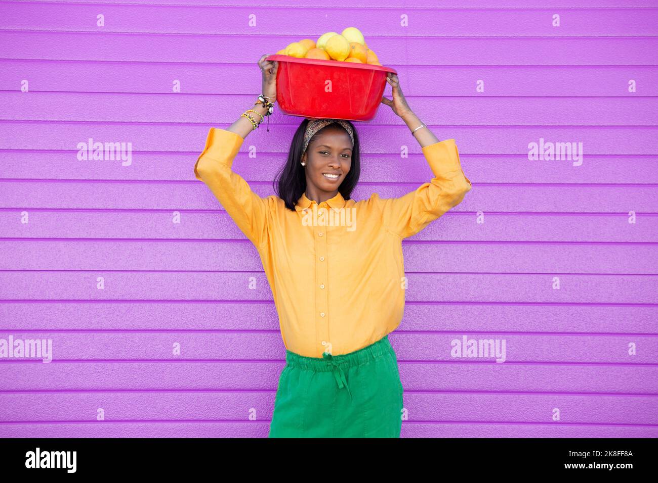 Young woman carrying basket full of oranges on head in front of purple wall Stock Photo