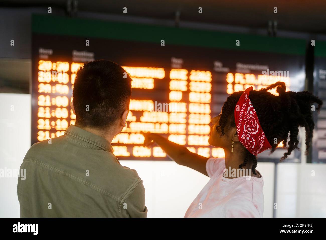 Woman with bandana pointing at departure board and talking to friend Stock Photo