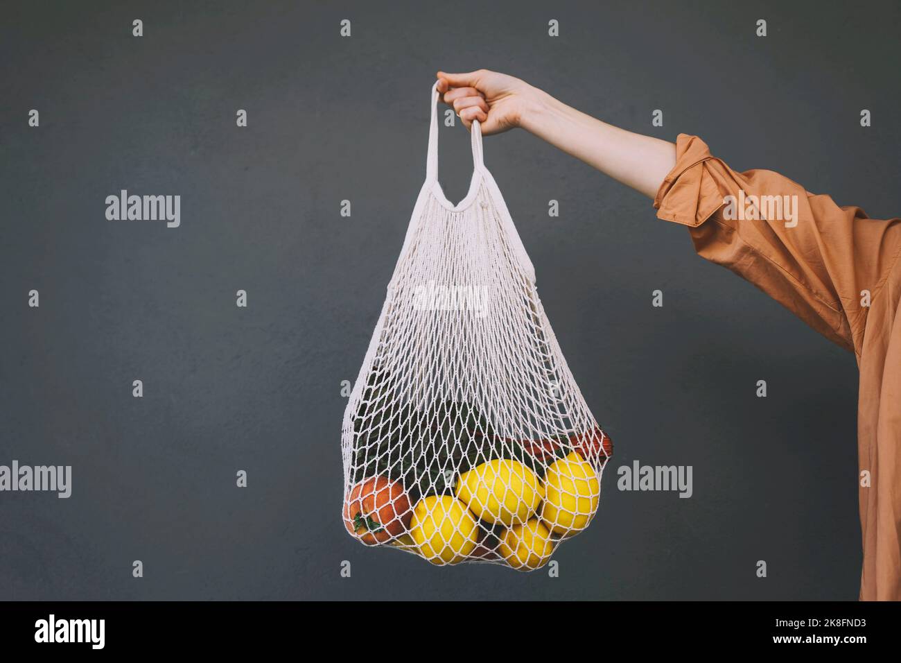 Woman holding lemons and vegetables in mesh bag against gray background Stock Photo