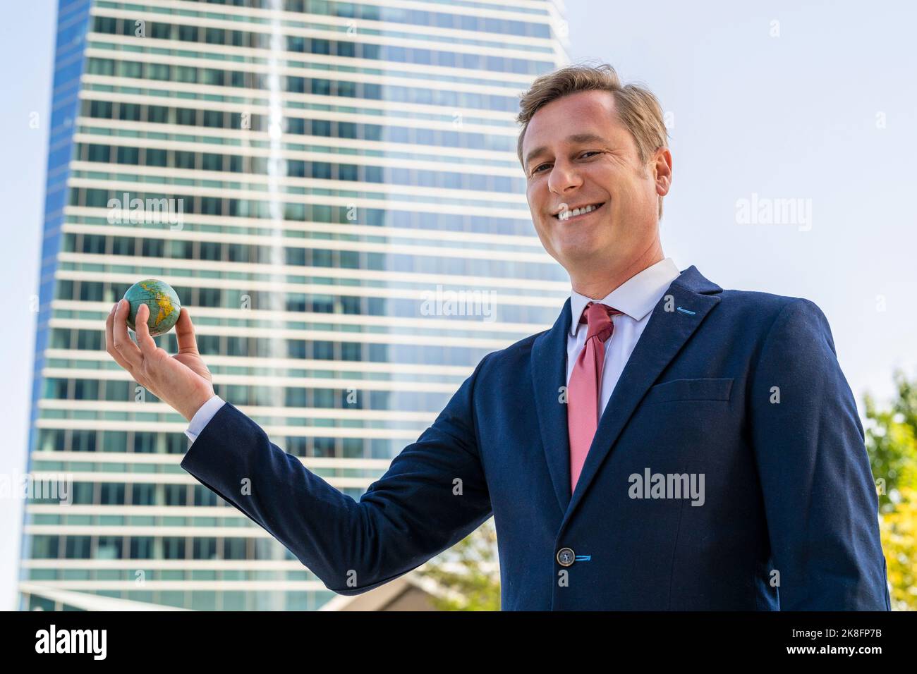 Smiling businessman holding small globe ball in front of office building Stock Photo