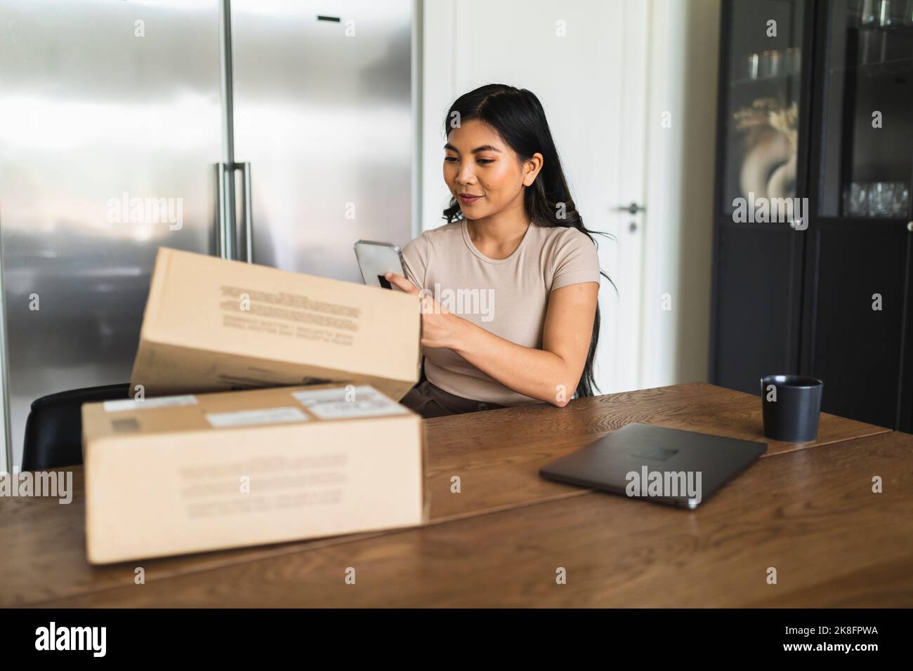 Young woman taking picture of delivery package box at dining table Stock Photo