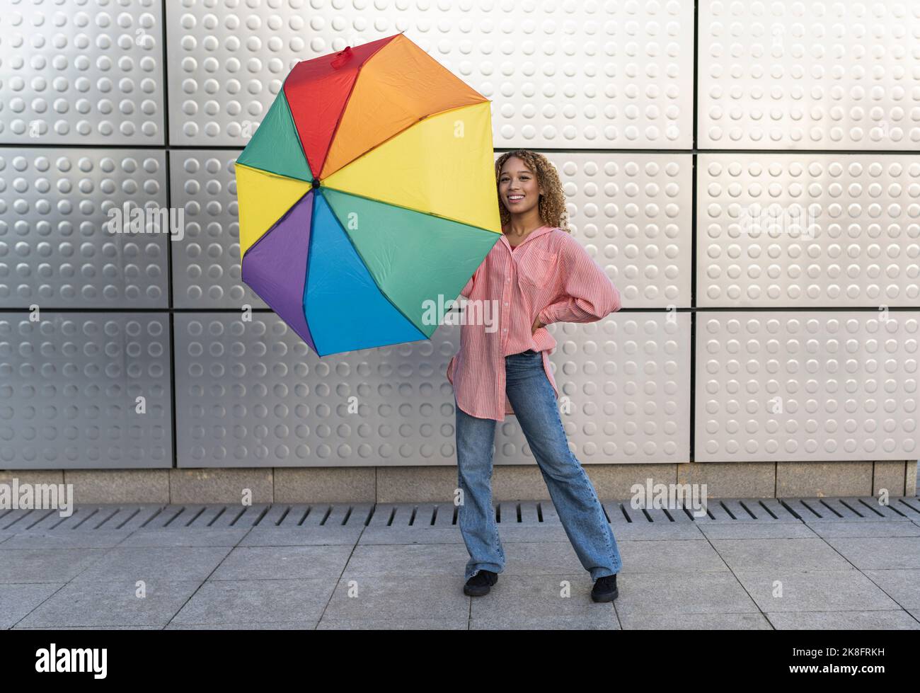 Smiling young woman with rainbow umbrella on footpath Stock Photo