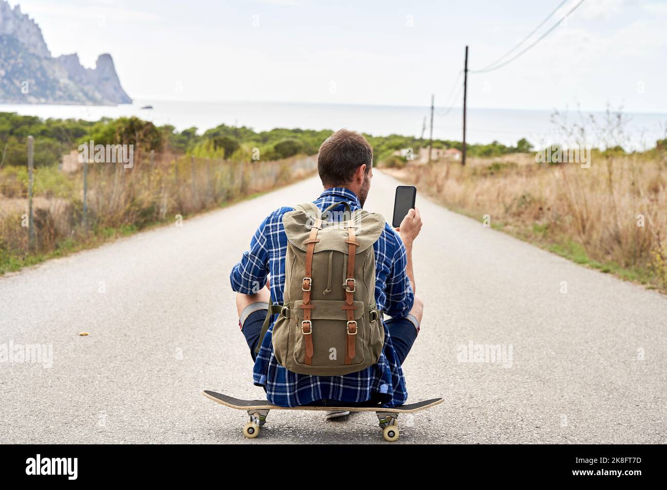 Man wearing backpack using smart phone sitting on skateboard Stock Photo
