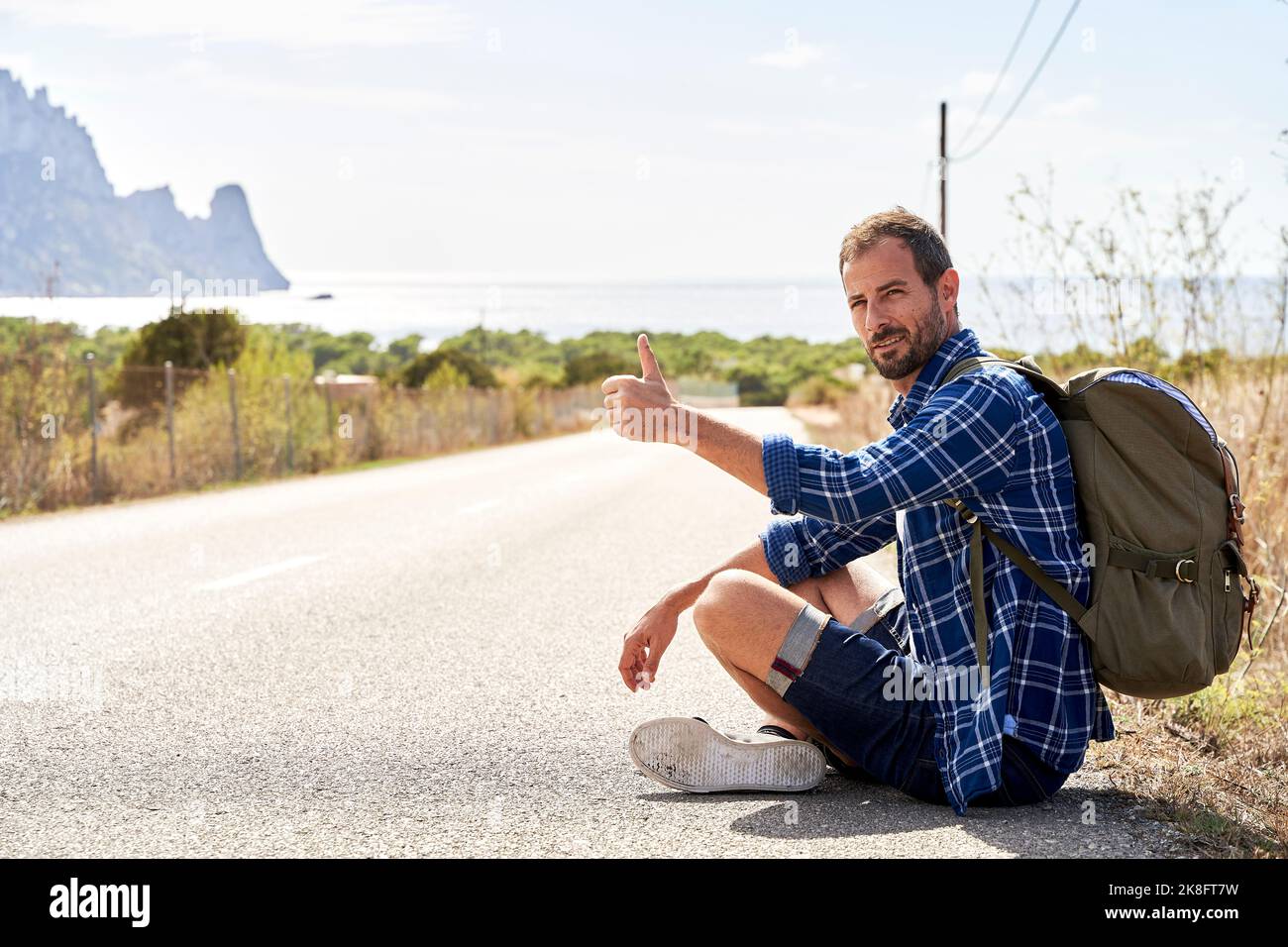 Man wearing backpack gesturing thumbs up sitting on road Stock Photo