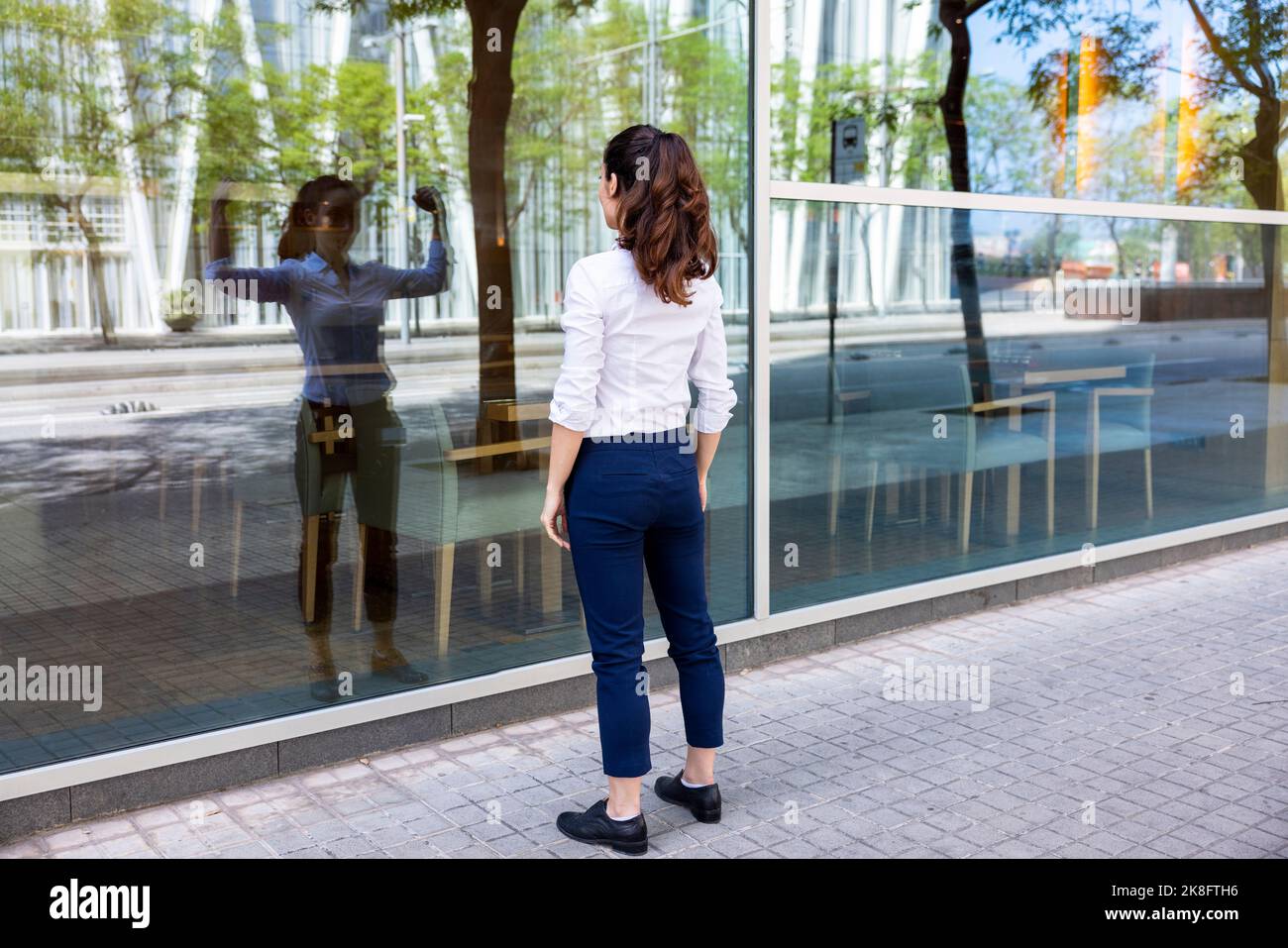 Businesswoman looking at reflection flexing muscles on glass wall Stock Photo