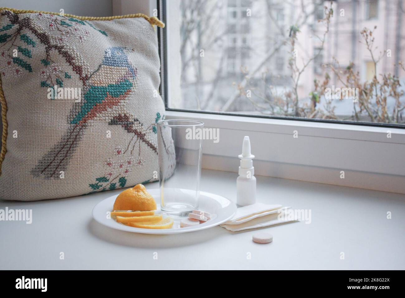 Seasonal cold: an empty glass cup, sliced lemon, instant pills and nasal spray with paper napkins on the windowsill. Next to the vintage embroidered p Stock Photo