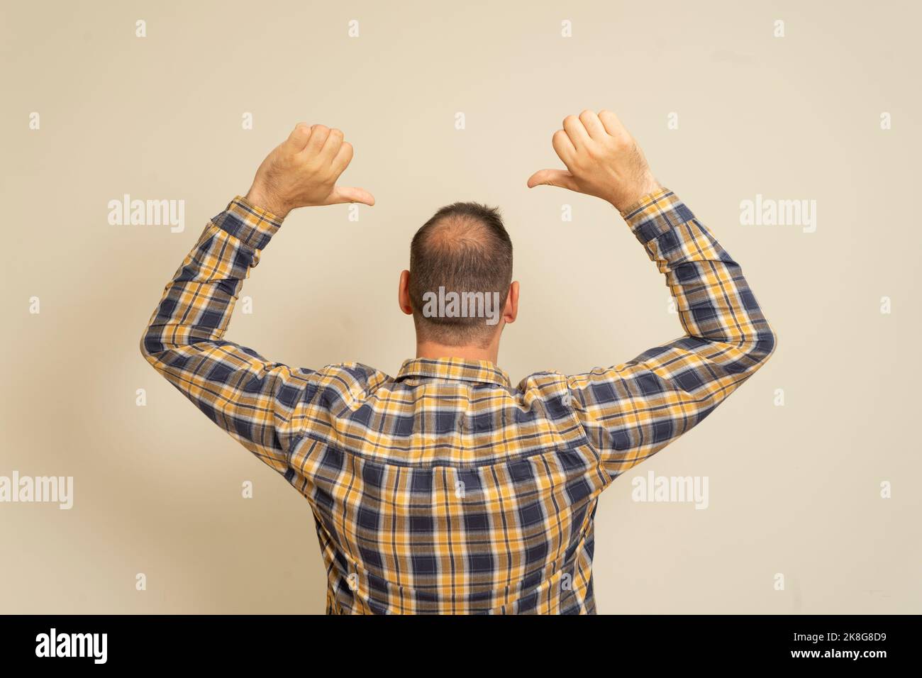 Portrait of a man with incipient alopecia from behind, raising both hands and pointing his index fingers upwards, isolated on a beige background. Stock Photo