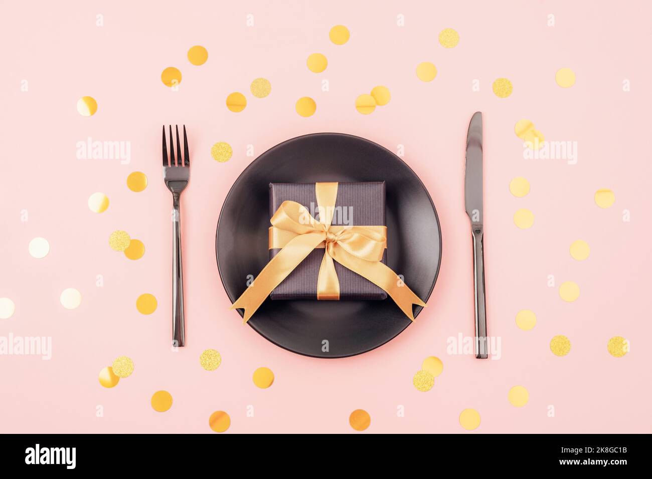Stylish table setting, gift box, black plate, cutlery on pink background with golden confetti. Christmas concept. Top view, flat lay Stock Photo