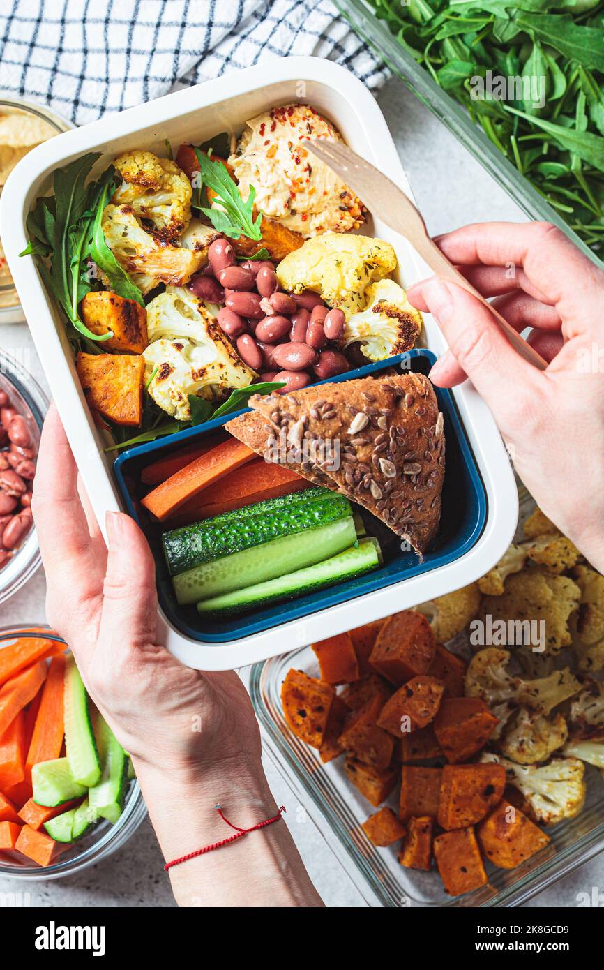 Cooking vegan lunch box flat lay. Hummus, beans, baked vegetables, green salad, rye bread and vegetable sticks in a plastic container, top view. Stock Photo