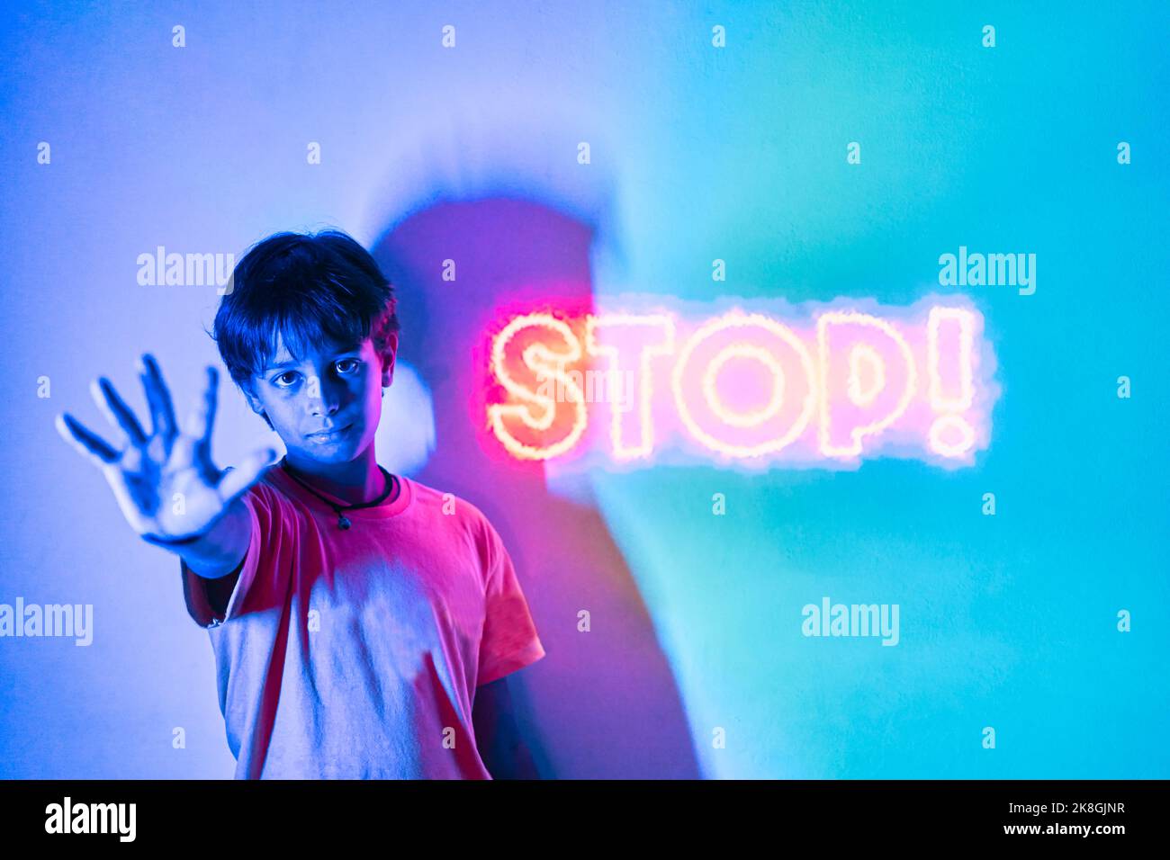 Determined preteen kid in t shirt standing in room with blue illumination and showing no gesture near orange neon stop inscription Stock Photo