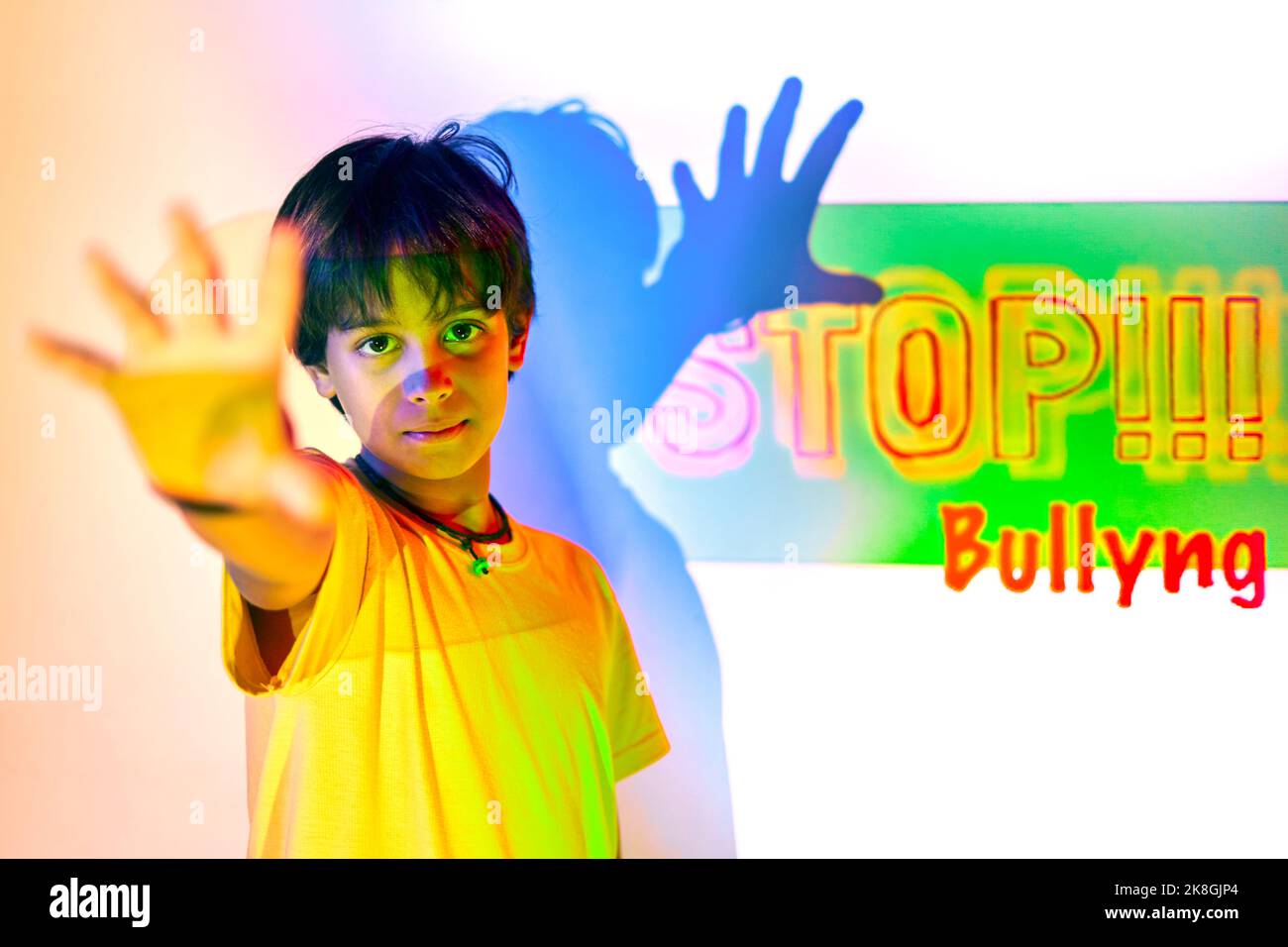 Serious Indian boy in yellow t shirt standing near white wall and outstretching arm showing no gesture against colorful neon stop bullying inscription Stock Photo