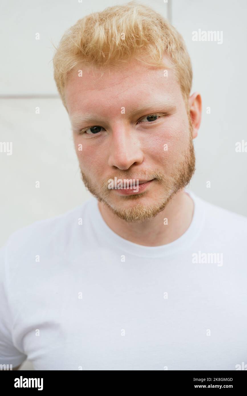 Pensive bearded adult blond haired man in white t shirt standing near white tiled wall looking at camera Stock Photo