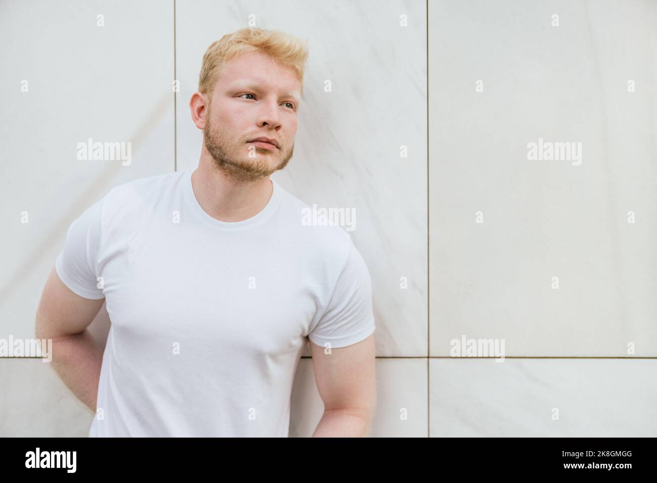 Pensive bearded adult blond haired man in white t shirt standing near white tiled wall looking away Stock Photo