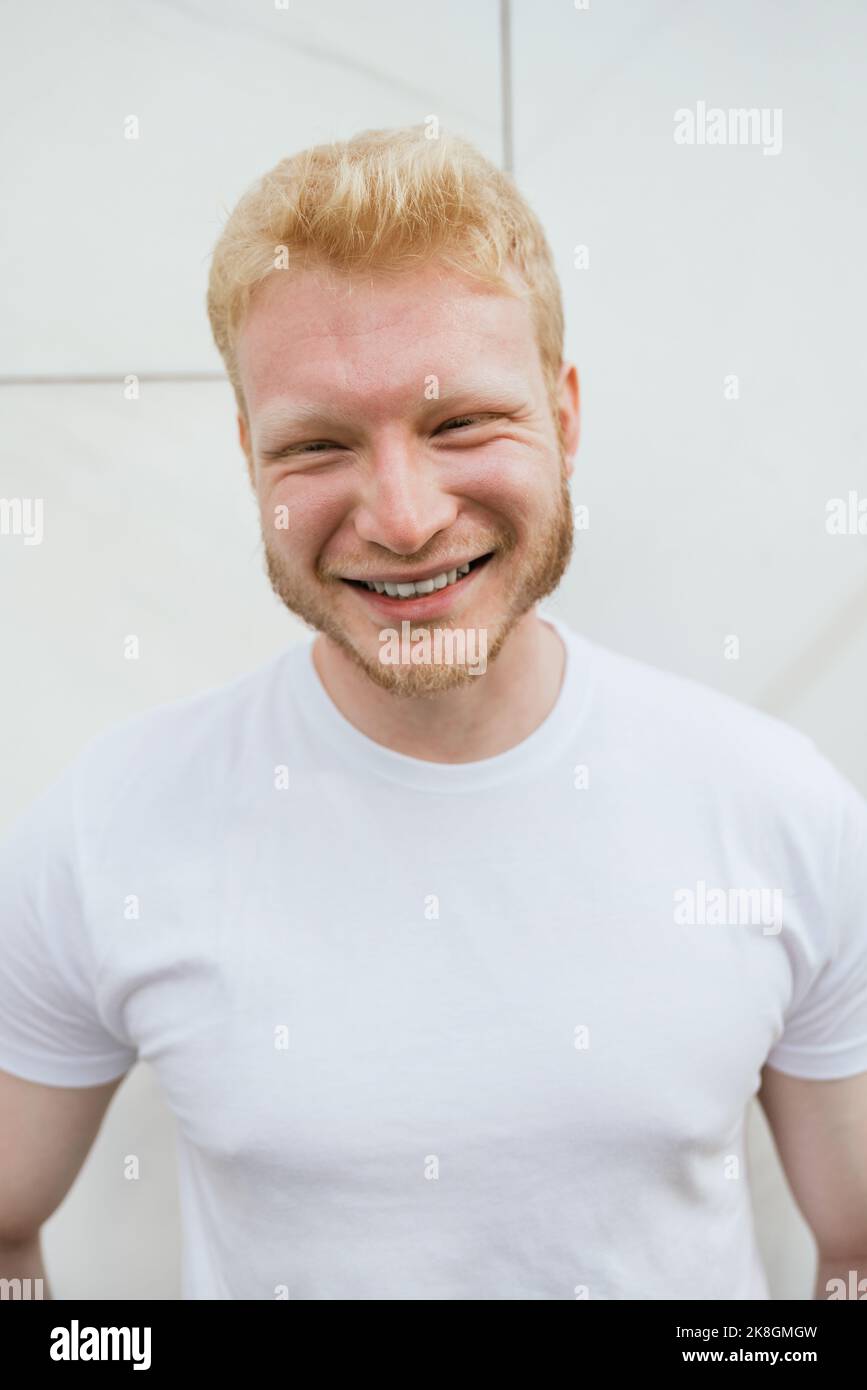 Cheerful bearded adult blond haired man in white t shirt standing near white tiled wall looking at camera Stock Photo