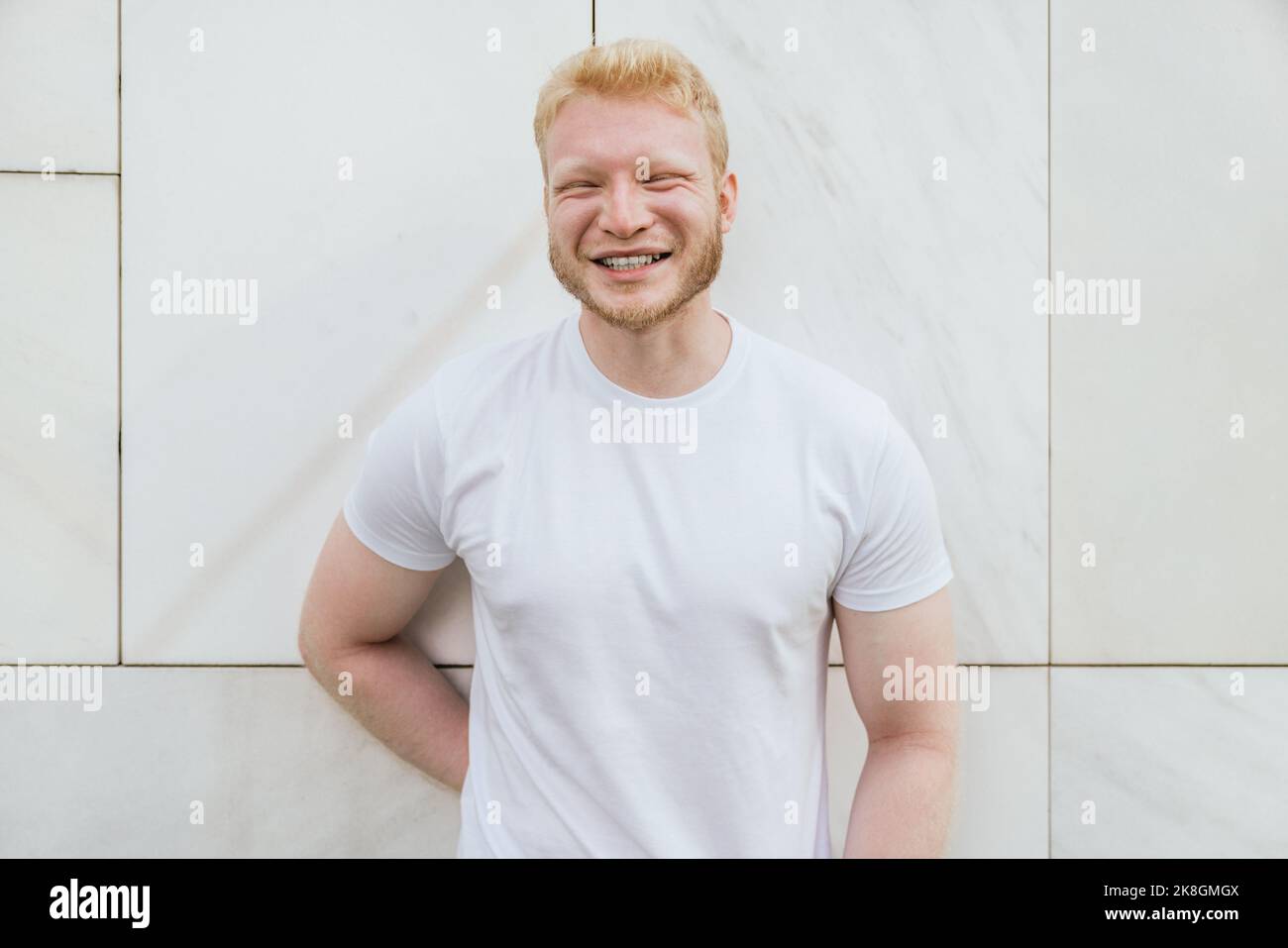 Cheerful bearded adult blond haired man in white t shirt standing near white tiled wall looking at camera Stock Photo