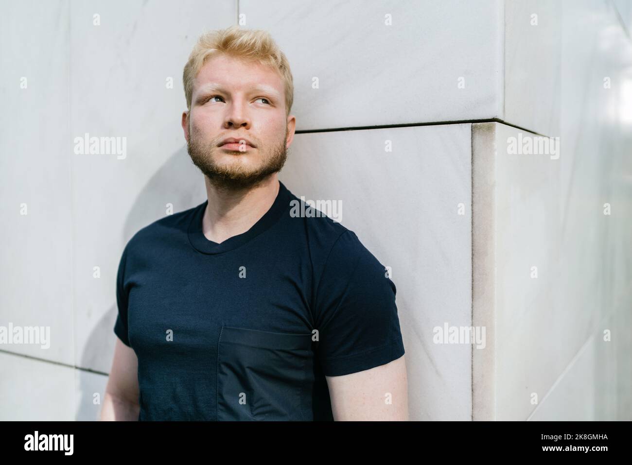Pensive bearded adult blond haired man in black t shirt standing near white tiled wall looking away Stock Photo