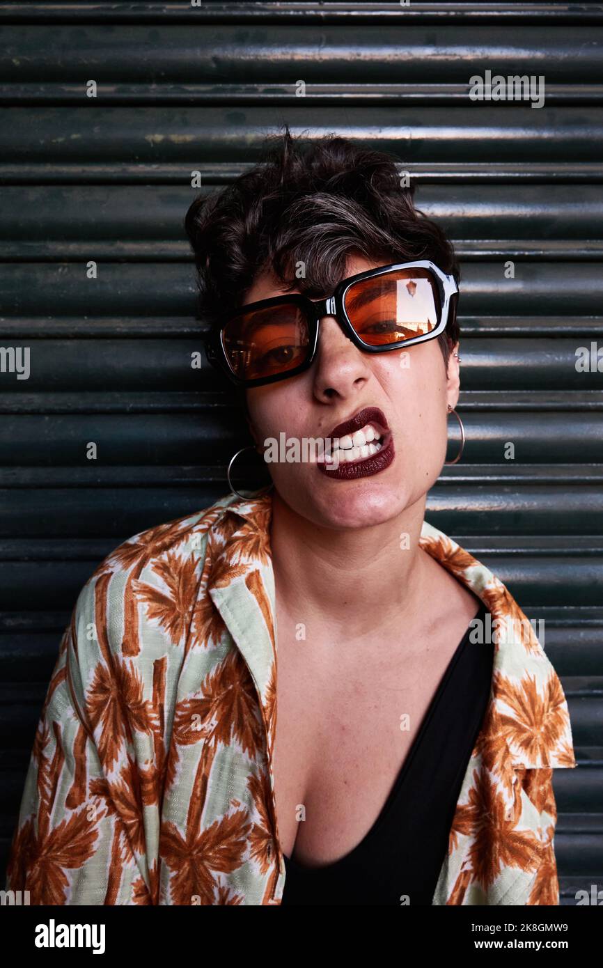 Stylish young woman in sunglasses with short hair sneering and looking at camera against black wall Stock Photo