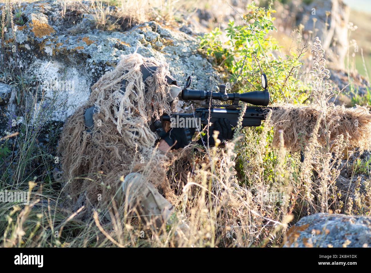 sniper in ghillie (camouflage) suit with a high-precision rifle with optic scope in a combat position in the mountains Stock Photo