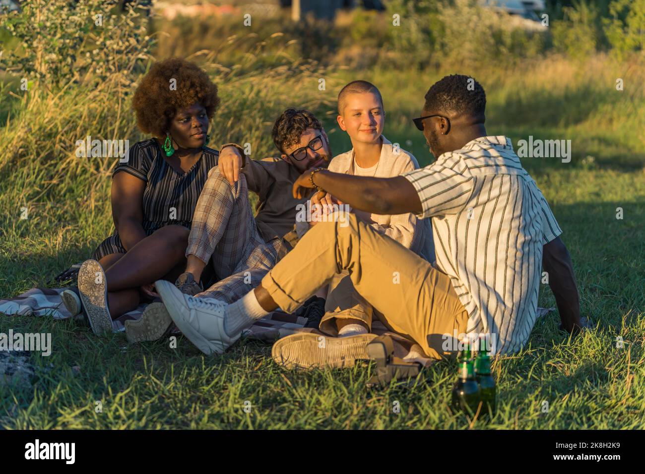 Outdoor party by the river. European youth. Multiethnic students in their mid 20s sitting on the grass, drinking beer, and talking. Meadow and golden hour sunshine. High quality photo Stock Photo