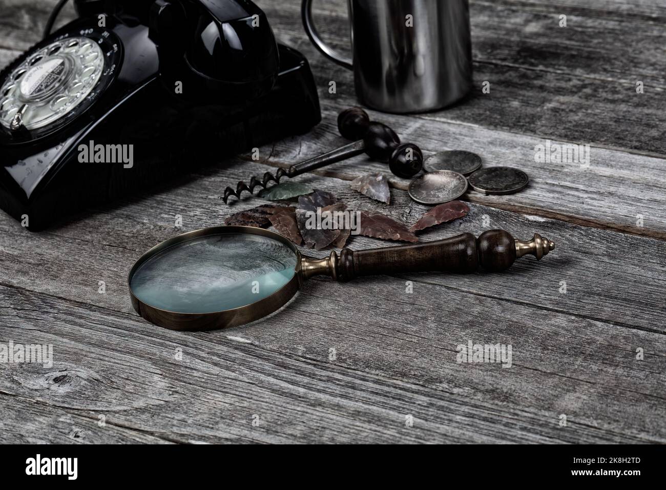 Vintage magnify glass, silver coins, native American arrowhead stones, wine corkscrew, steel coffee cup, and rotary phone on rustic wood table for col Stock Photo