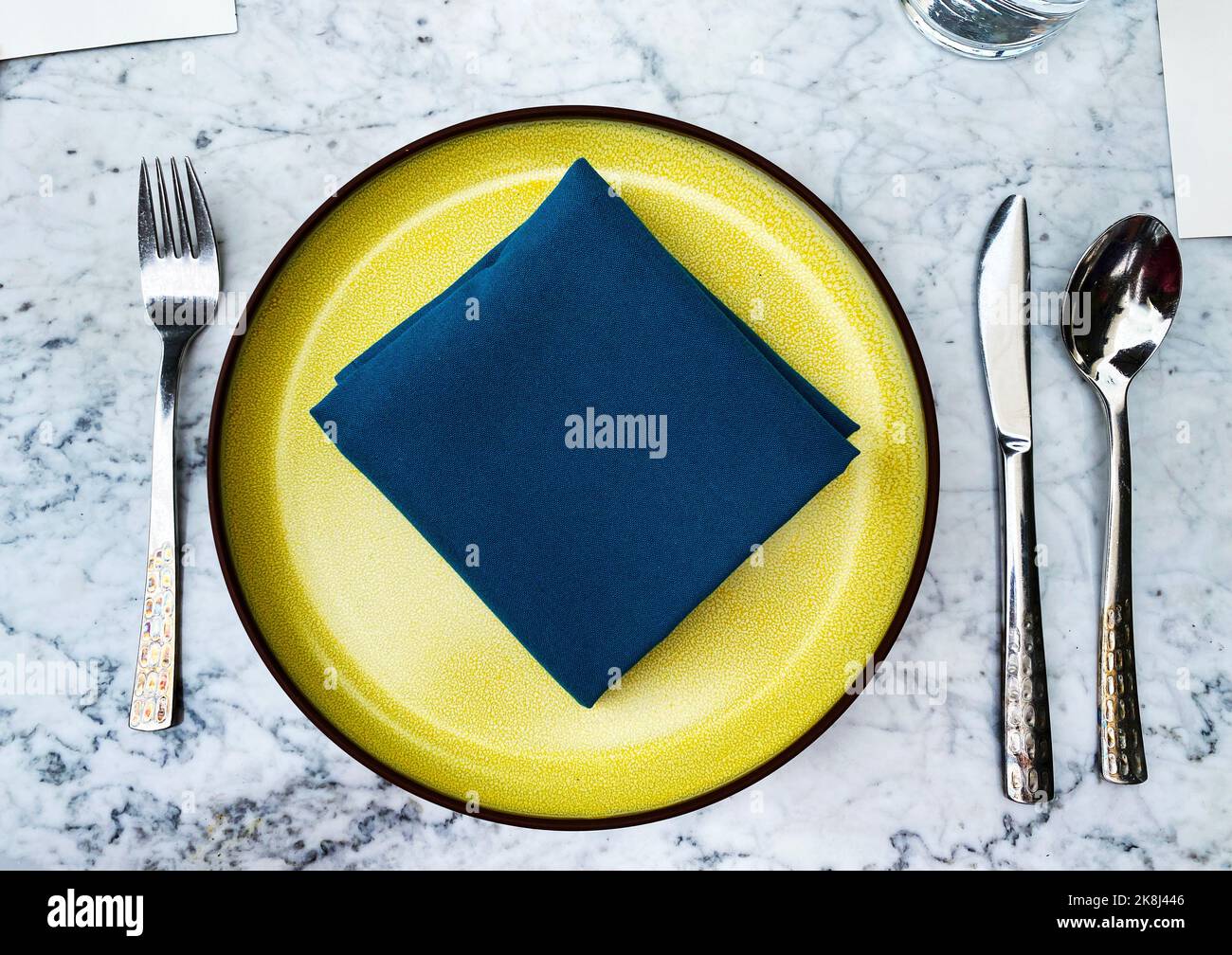 A top down flat lay of a place setting with a blue napkin on a yellow plate atop a marble table. Stock Photo