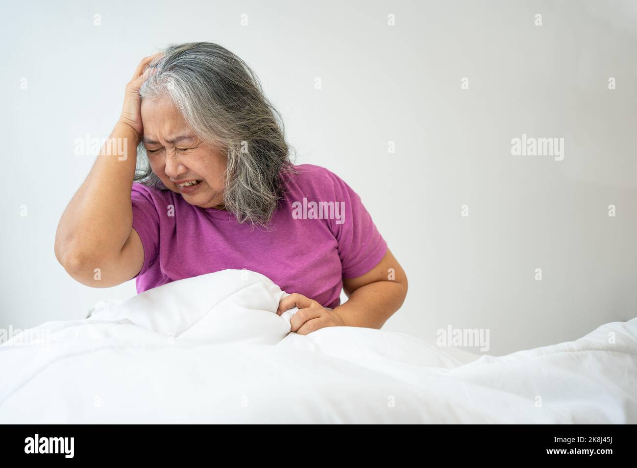 Unhealthy elderly senior Asian woman sitting on bed suffering from headache and migraine so pain and illness, Stress senior woman sit on bed at home a Stock Photo