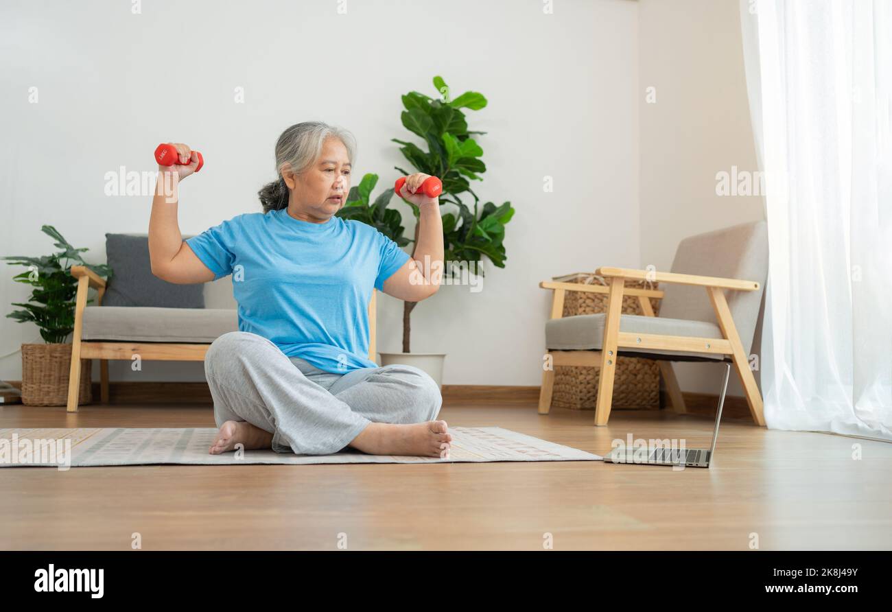 Asian senior woman lifting dumbbell for exercise and workout at home. Active mature woman doing stretching exercise in living room. Exercise Active an Stock Photo