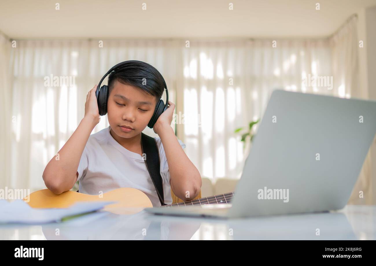 Asian boy playing guitar and watching online course on laptop while practicing for learning music or musical instrument online at home. Boy students s Stock Photo