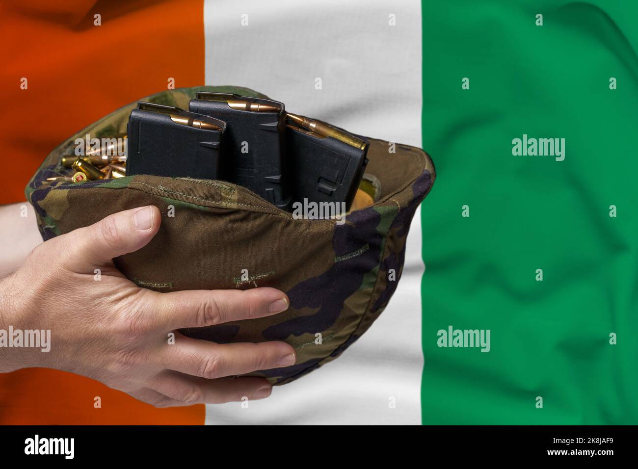 A military helmet with cartridges and magazines for a rifle in the hands of a man against the background of the flag of Ivory Coast. The concept of Stock Photo