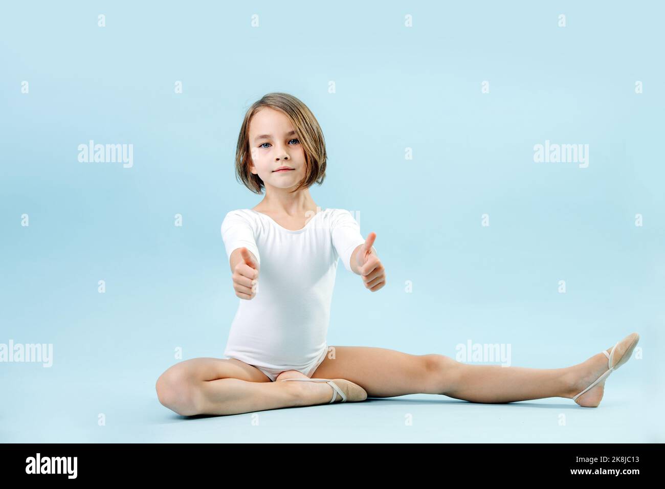 Tired girl in a white leotard sitting in half splits, giving thumbs up, looking at the camera. Over blue background. Stock Photo