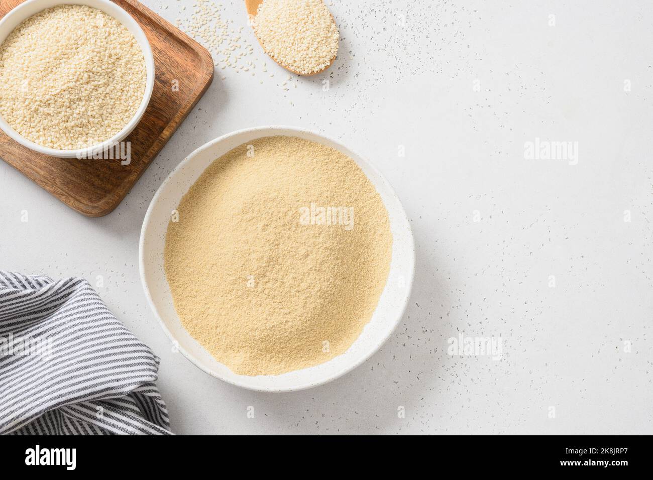 Sesame flour in bowl and white sesame seeds in spoon on white background. View from above. Copy space. Stock Photo