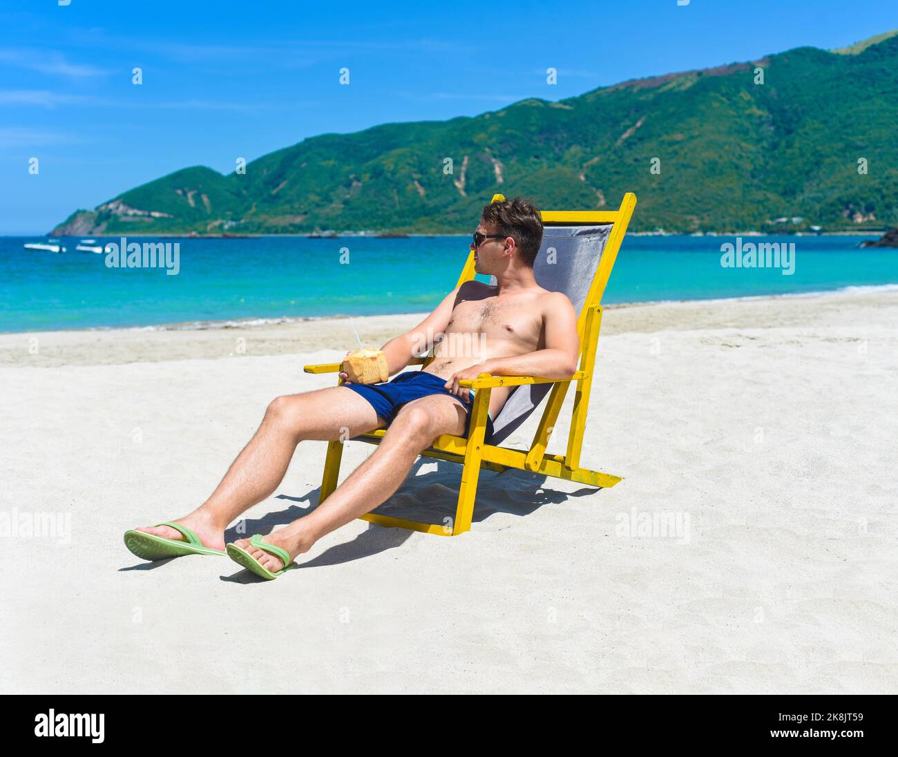 Young happy man drinks coconut juice sitting on deck chair on a south china sea beach in vietnam Stock Photo