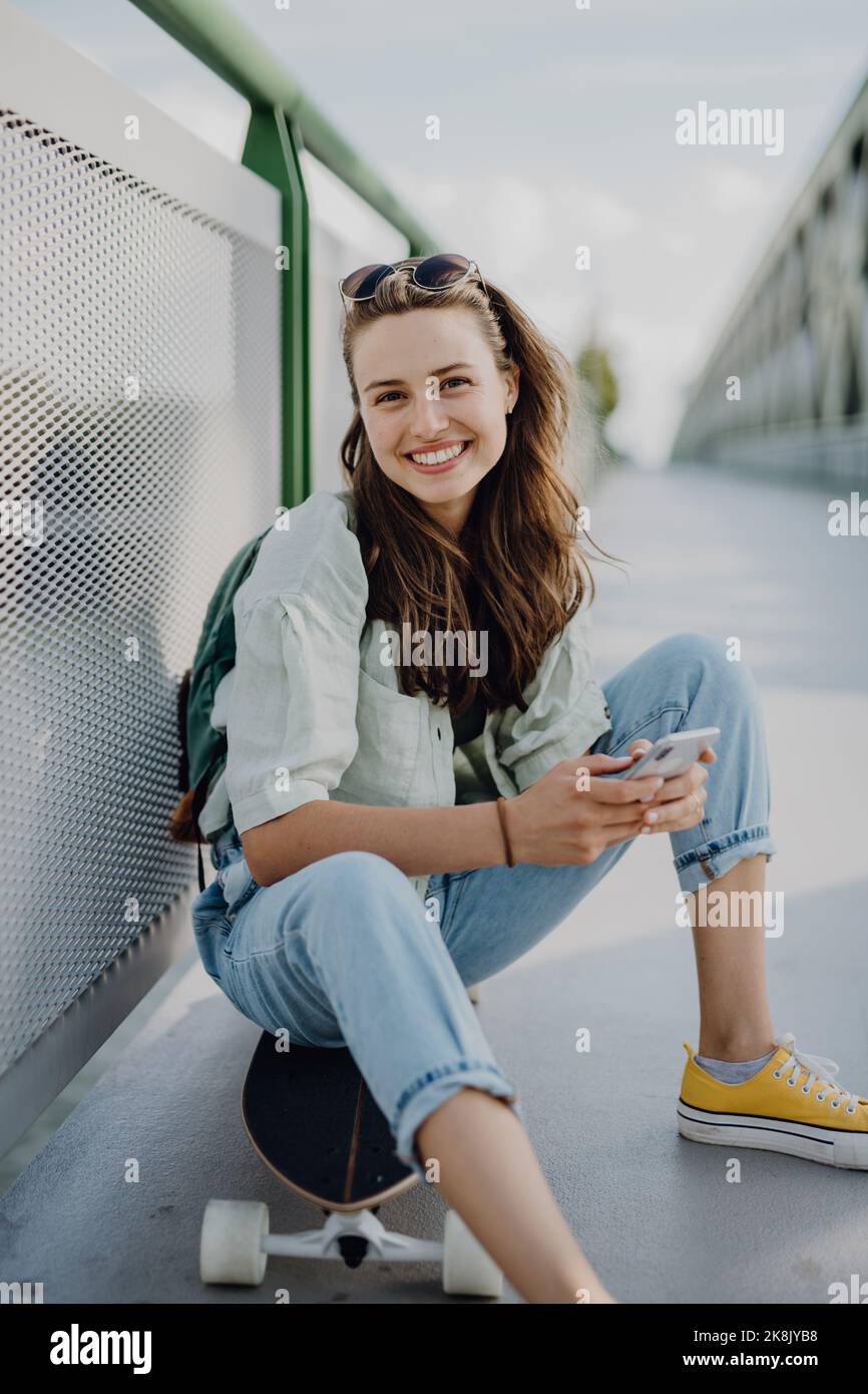 Happy young woman sitting on skateboard at city bridge. Youth culture and commuting concept. Stock Photo