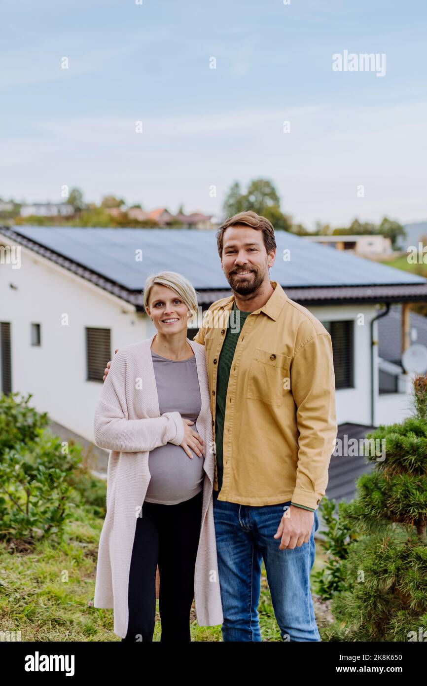 Happy man with his pregnant wife standing in front of their new house with photovoltaics solar panels. Stock Photo