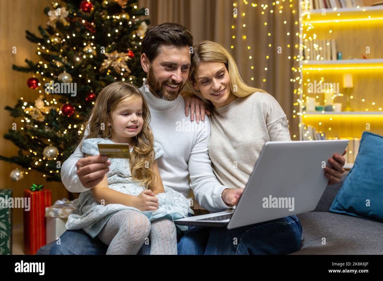 Christmas family, husband wife and little daughter for Christmas at home make online shopping in online store remotely, smiling and happy, husband holding bank credit card and laptop choosing gifts. Stock Photo