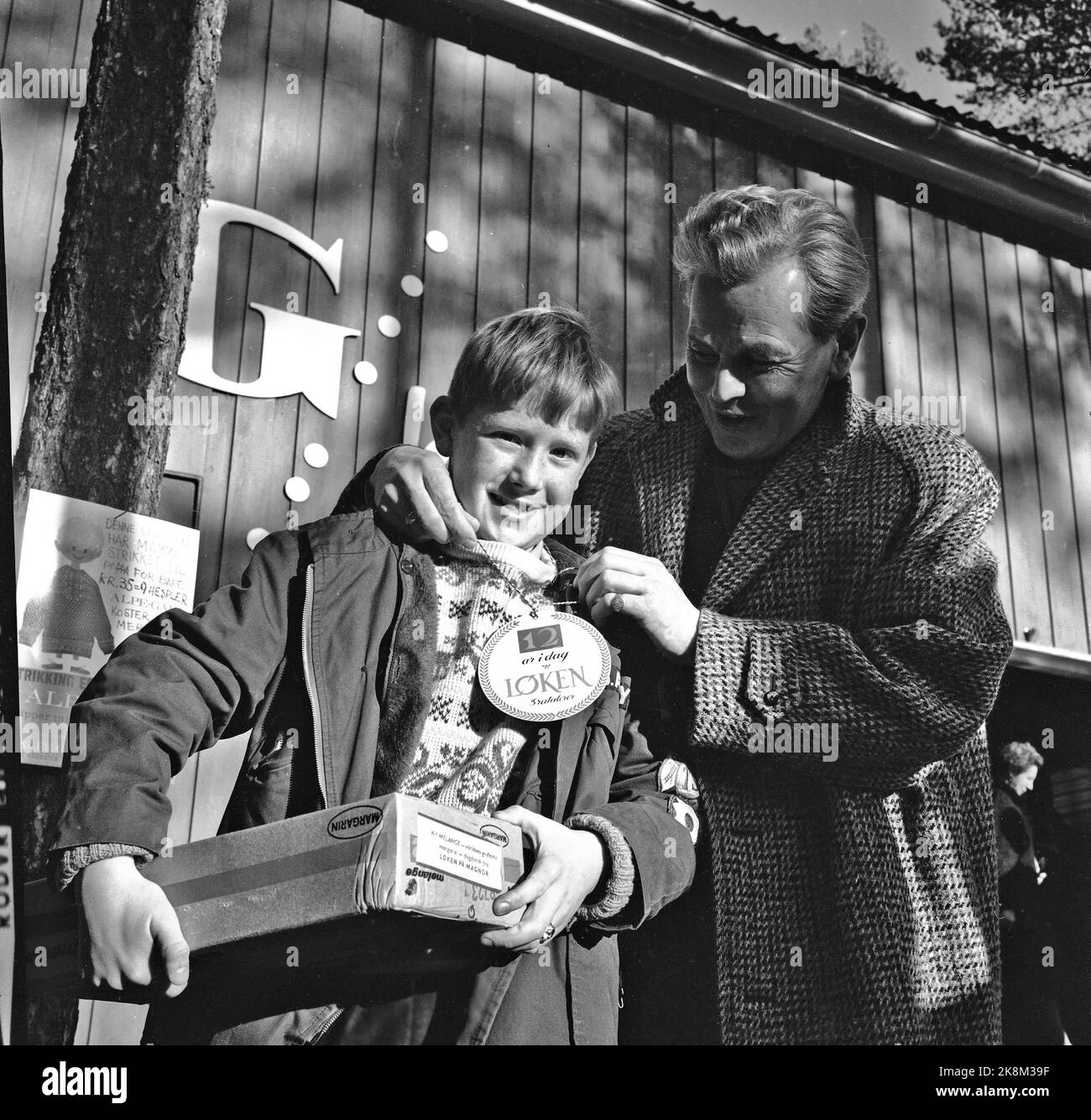 Magnor oktober 1965: KASSASUKSESS FOR NORSK MARGARIN Allehelgensdag kom nærmere ti tusen stappfulle biler fra Sverige til Magnor for å handle subsidierte norske matvarer som sukker, margarin og hvetemel. De aller fleste handlet i varehuset til 'margarinkongen' Per Løken. Her overrekker han gave til 12 år gamle Bo. Barn som fylte 12 år den dagen fikk 5 kg margarin i gave. De var da 'Margarinmodne' (evt Sukkerunger) med rett til å ta med egen kvote av de dyrebare varene. Flere biler hadde med seg lånte unger i denne anledning.  Foto: Aage Storløkken / Aktuell / NTB Stock Photo