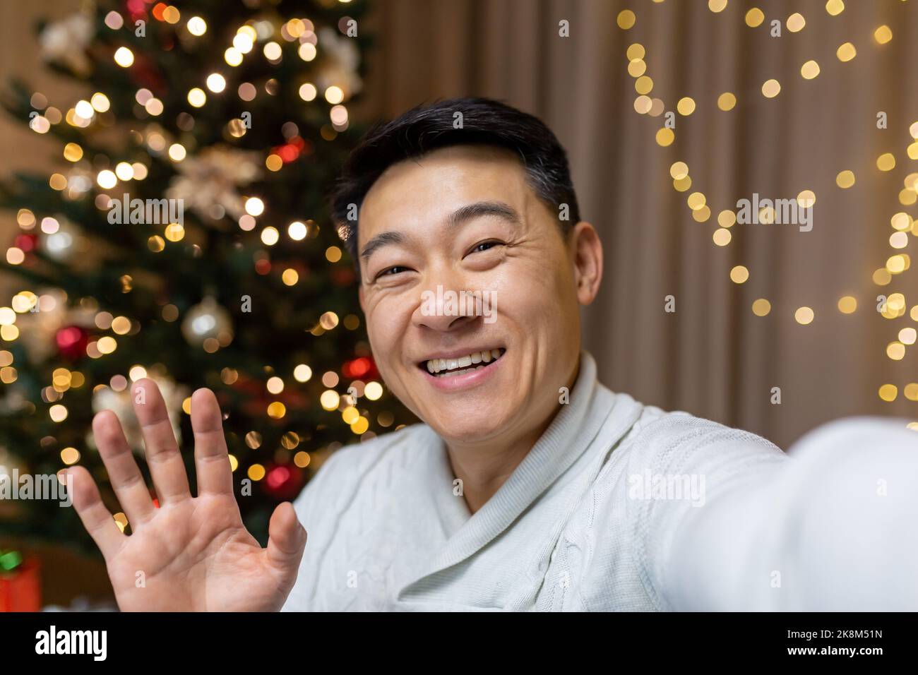 Happy asian man celebrating christmas and new year alone at home, man looking at smartphone camera and smiling, talking with friends on phone video call inviting guests while sitting on sofa Stock Photo