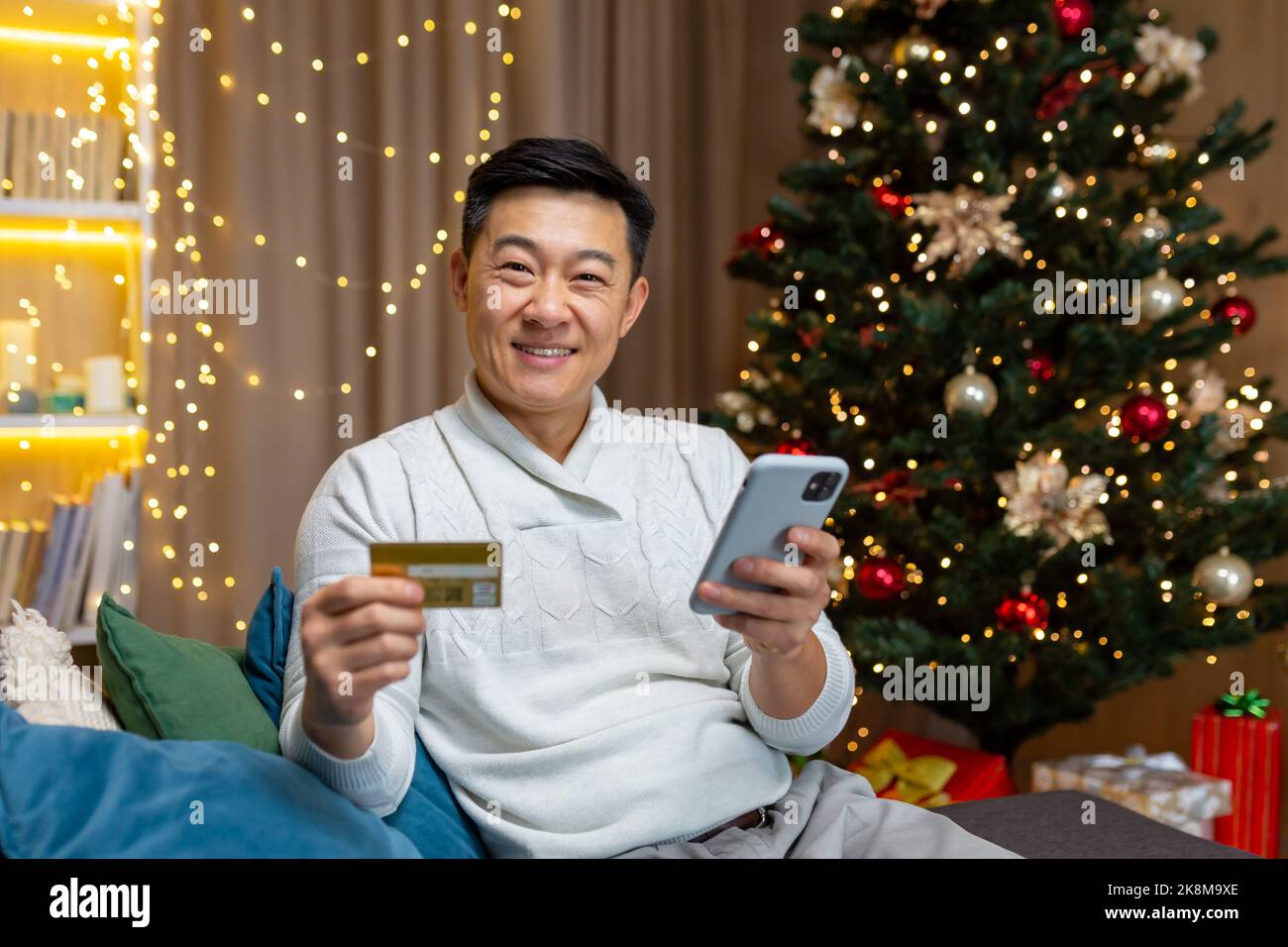 Happy Asian online shopping on new year Christmas eve buying and choosing gifts for friends in online store, portrait of happy man home looking at camera and smiling with bank credit card and phone. Stock Photo