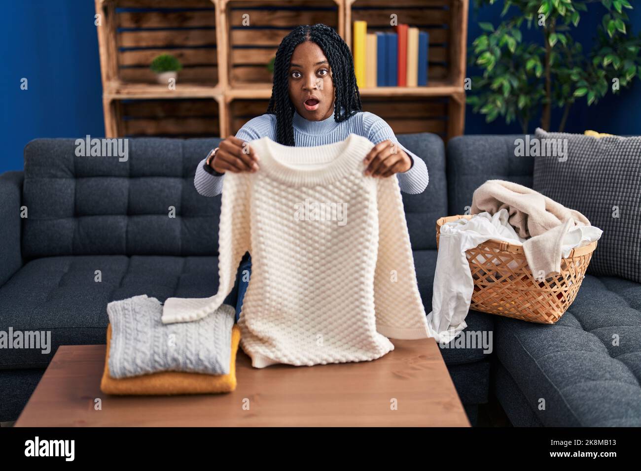 African american woman folding clean laundry in shock face, looking
